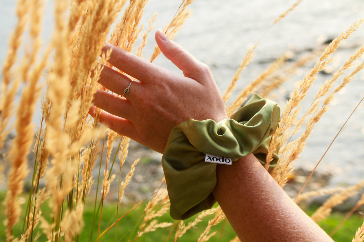 Hand wearing a green scrunchie with a brand logo, touching tall grass.