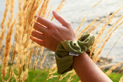 Hand wearing a green scrunchie with a brand logo, touching tall grass.